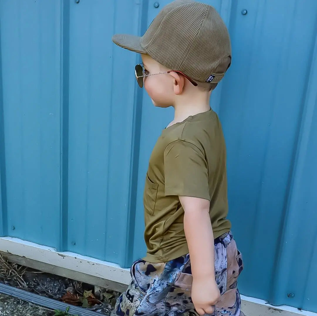 Little boy wearing an Olive Bamboo Boys Shirt and cap, enjoying a sunny day outdoors.
