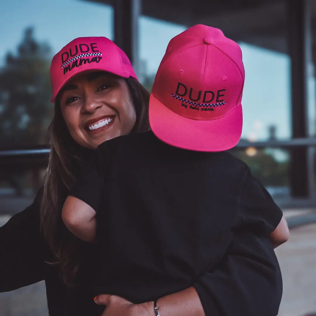 Smiling woman wearing a hot pink hat with a toddler in a matching hot pink toddler hat outdoors.