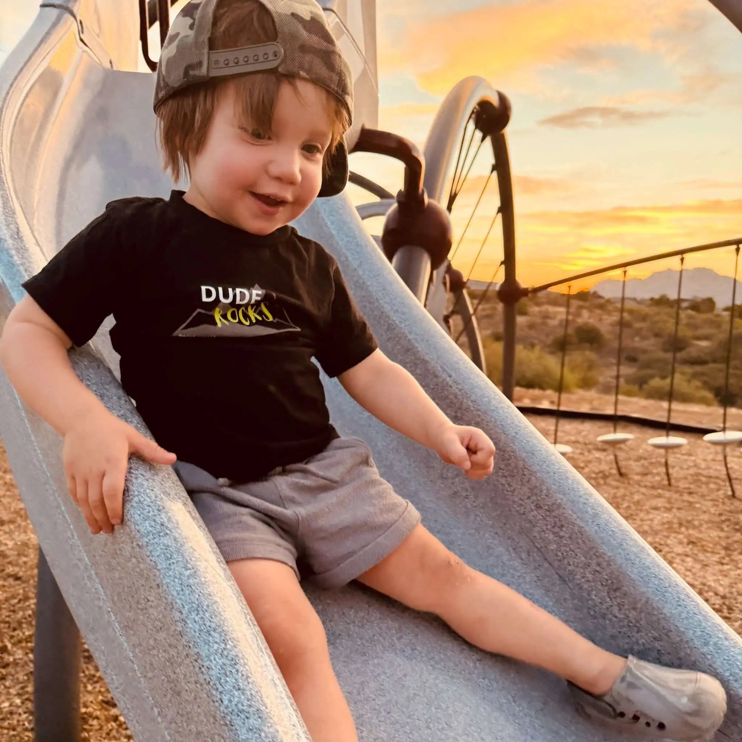 Young boy sliding down a playground slide wearing a Dude Rocks Shirt, showcasing fun and adventure in outdoor play.