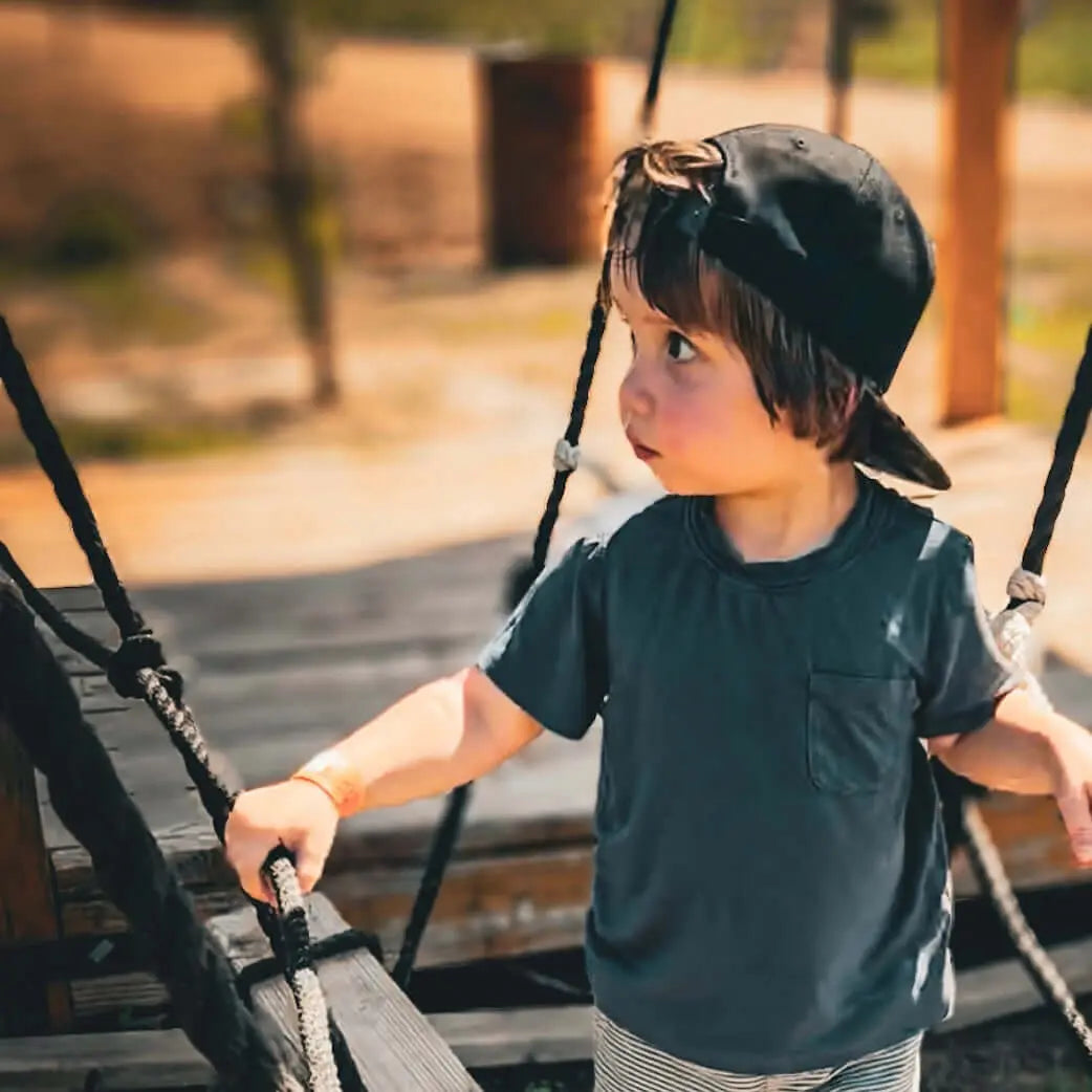 Young boy playing on a swing wearing a blue bamboo boys shirt and a cap.