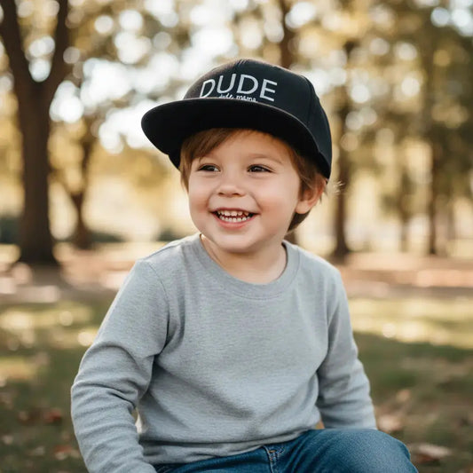 Little boy outside smiling wearing a kids hat that says 'Dude'.