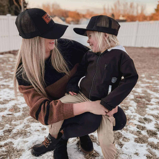 Mom and son are smiling at each other wearing matching black mom and son hats.