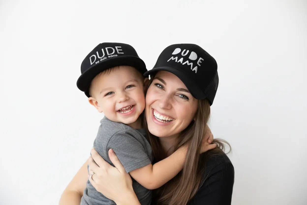 Mom and son are wearing matching hats - both hats are black with a matching gray font. The mama hat says 'Dude Mama' and the boys hat says 'Dude'.