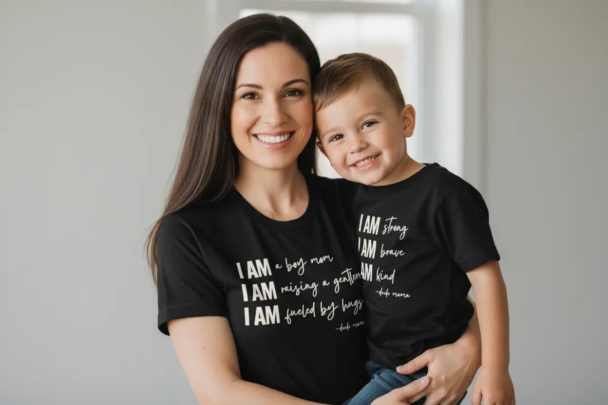 A mom holding her son. You can feel the love in this photo - they are wearing mom and son matching black shirts. It is an adorable mom and son matching outfit. The text on both shirts is off white. Mama's shirt says 'I am a boy mom, I am raising a gentleman, I am fueled by hugs.' Her son's shirt says 'I am strong, I am brave, I am kind.'