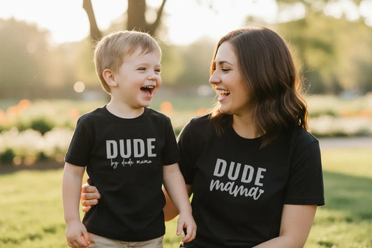 Mom and son wearing a matching outfit. Mama's black shirt says 'Dude Mama' in gray text and her son's black shirt says 'Dude' in gray text. They are happy and having fun outside.