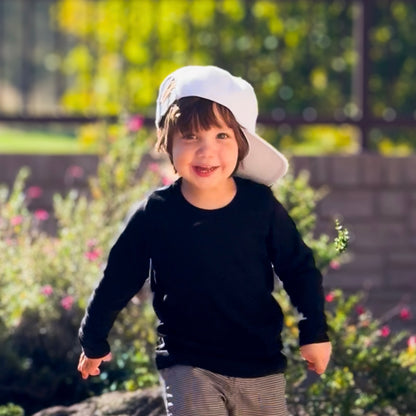 Smiling boy wearing a Dude Gray on White boy hat, running in a sunny garden.