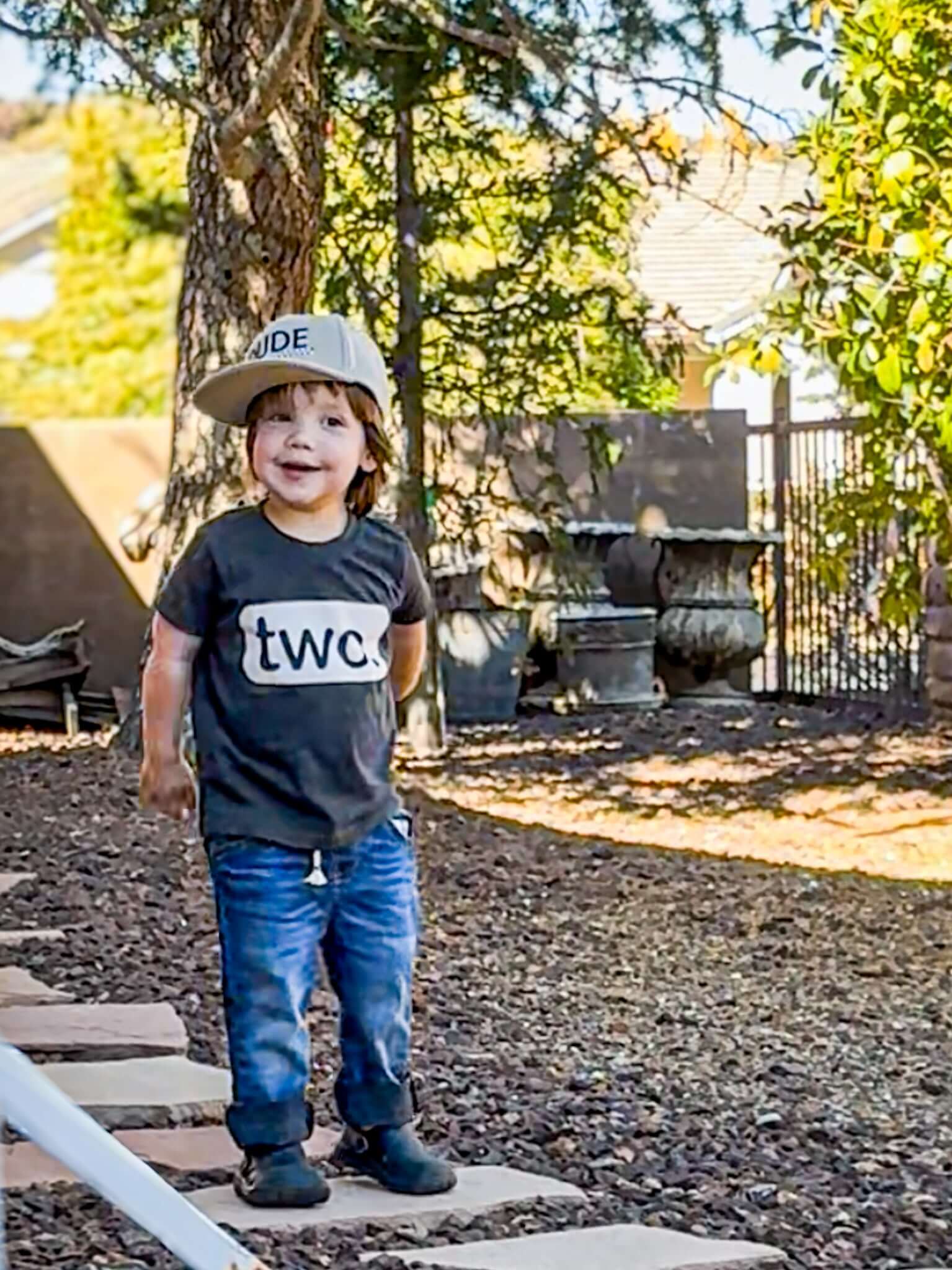 Boy wearing a khaki and checkered dude boy hat, smiling outdoors in a garden setting.