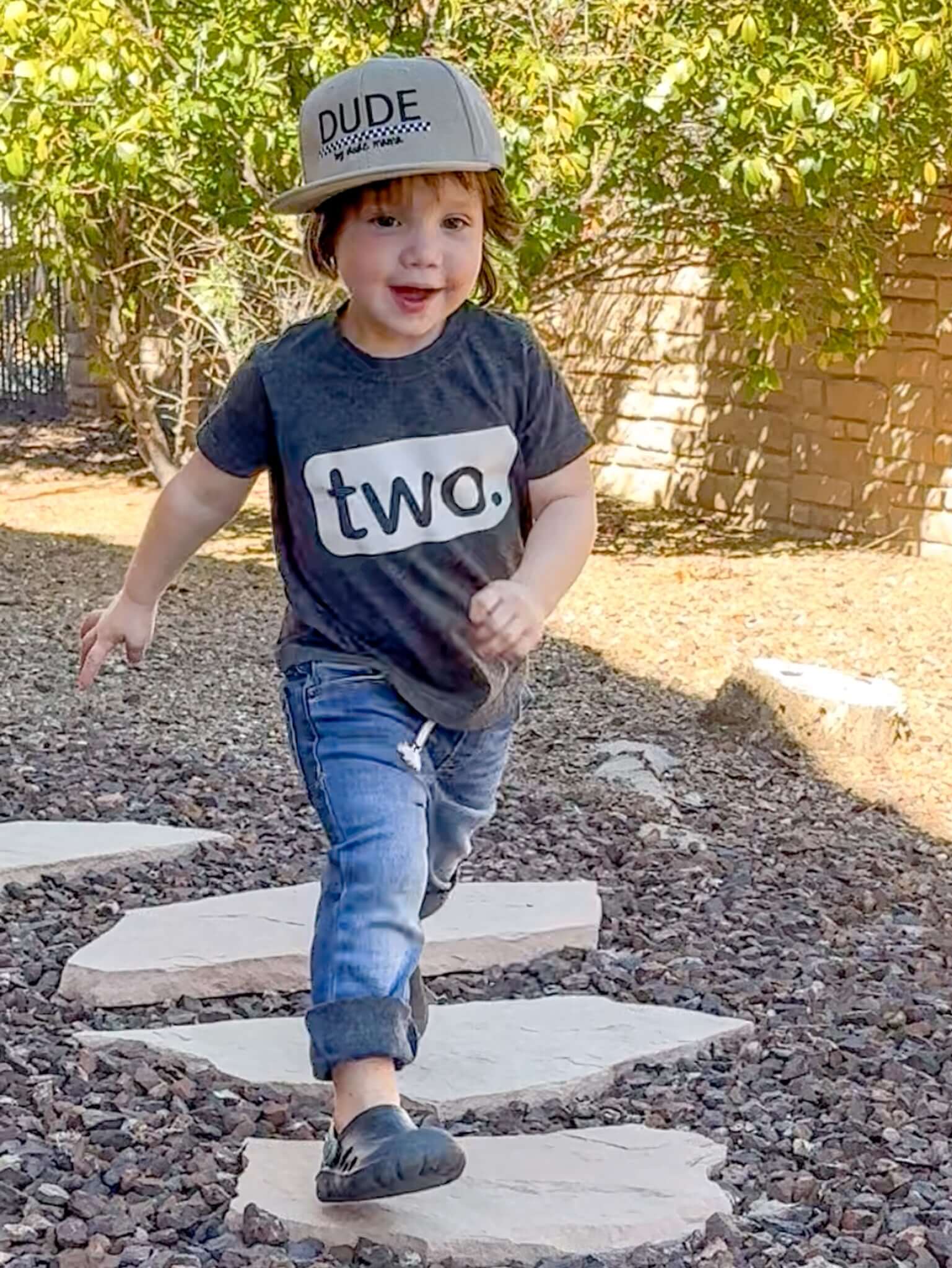 Joyful boy wearing khaki and checkered dude boy hat, running along stepping stones in casual outfit.