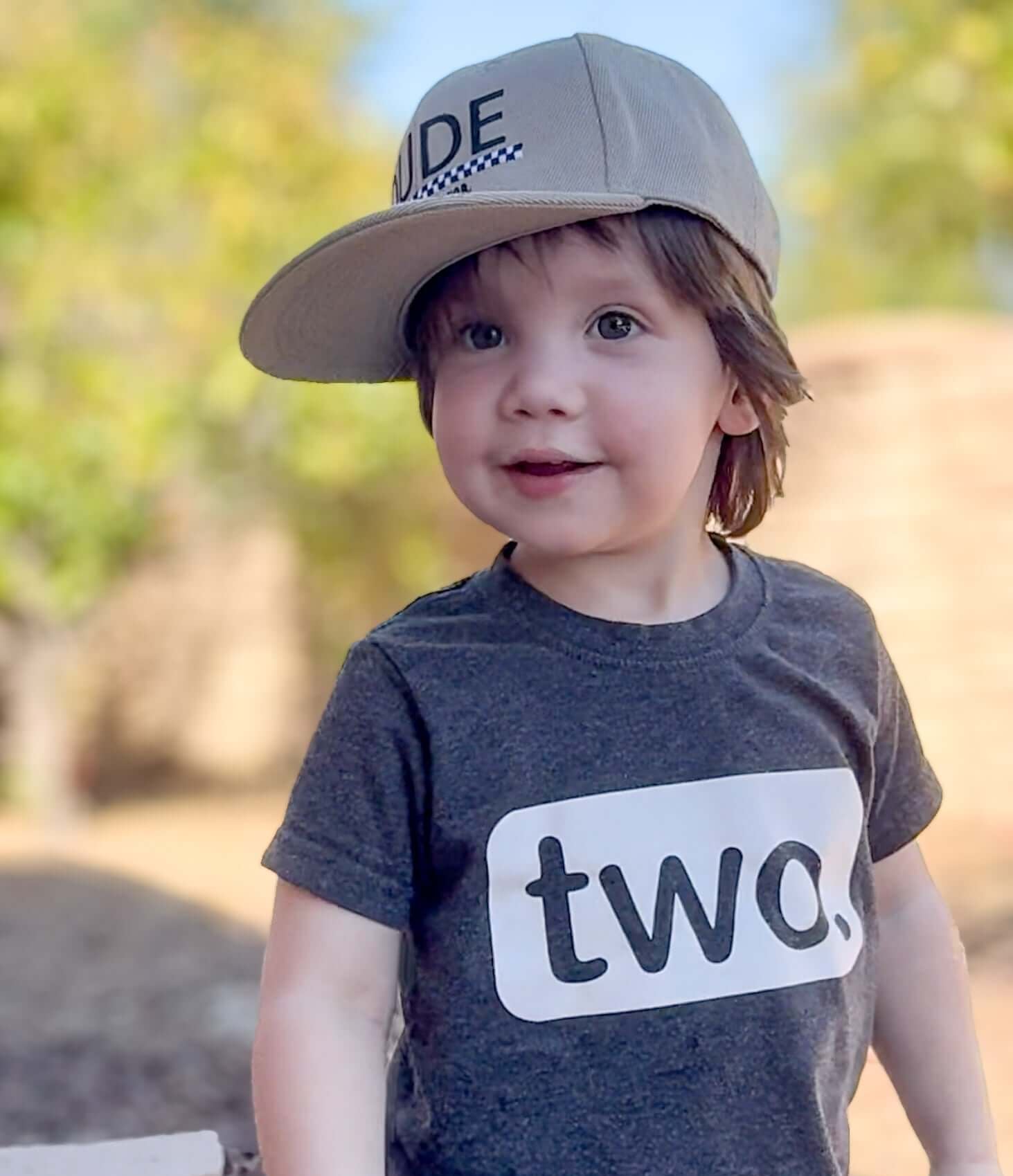 Boy wearing a khaki and checkered Dude boy hat, smiling confidently in a gray t-shirt that says 'two'.