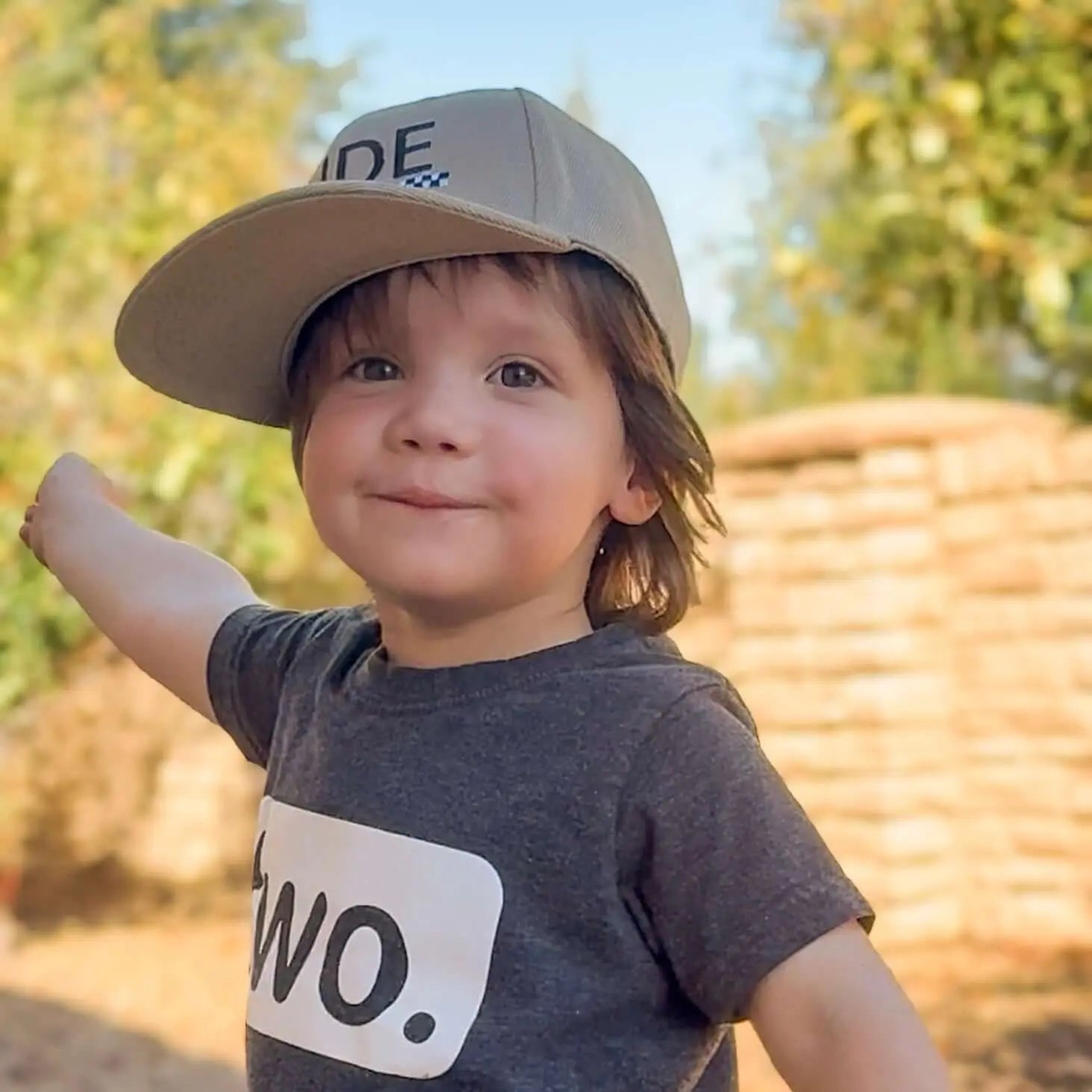 A young boy wearing a khaki & checkered dude boy hat, smiling confidently outdoors.
