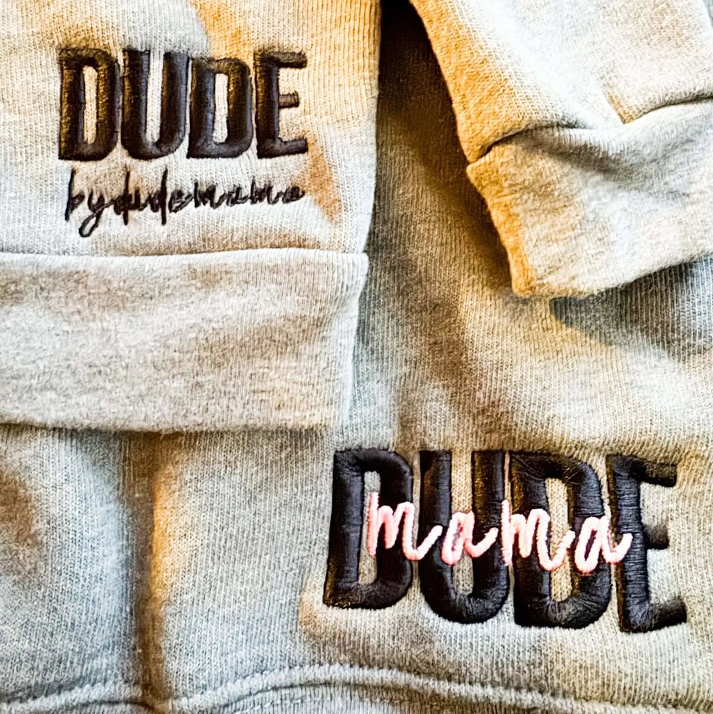 Mom and son matching outfit. It is two gray sweatshirts one for the boy and one for mom. The boys sweatshirt says "Dude" and the moms sweatshirt says "Dude Mama". The picture is zoomed in on the two sweatshirts showing the text.