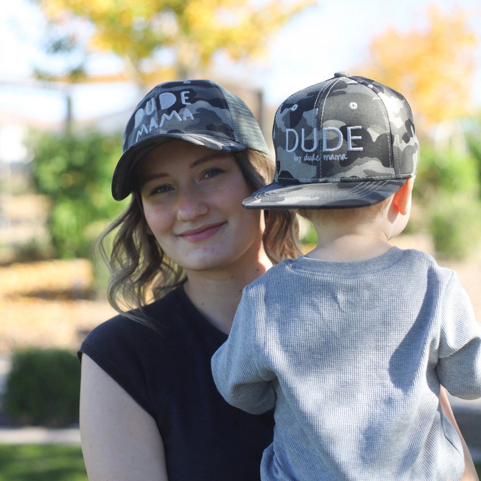 A boy mom wearing a Camo + Gray Hat that says 'Dude Mama' while holding her boy outdoors. Her little boy is wearing a hat that says 'Dude'. They are wearing matching mom and son hats.