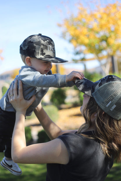 Matching mom and son hats being worn outdoors, with a mother lifting her son in the air, both wearing camo hats.