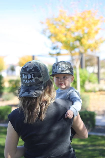 Matching mom and son hats featuring camo and gray design, with 'Dude' on the boy hat and 'Dude Mama' on the boy mom hat in a sunny park.