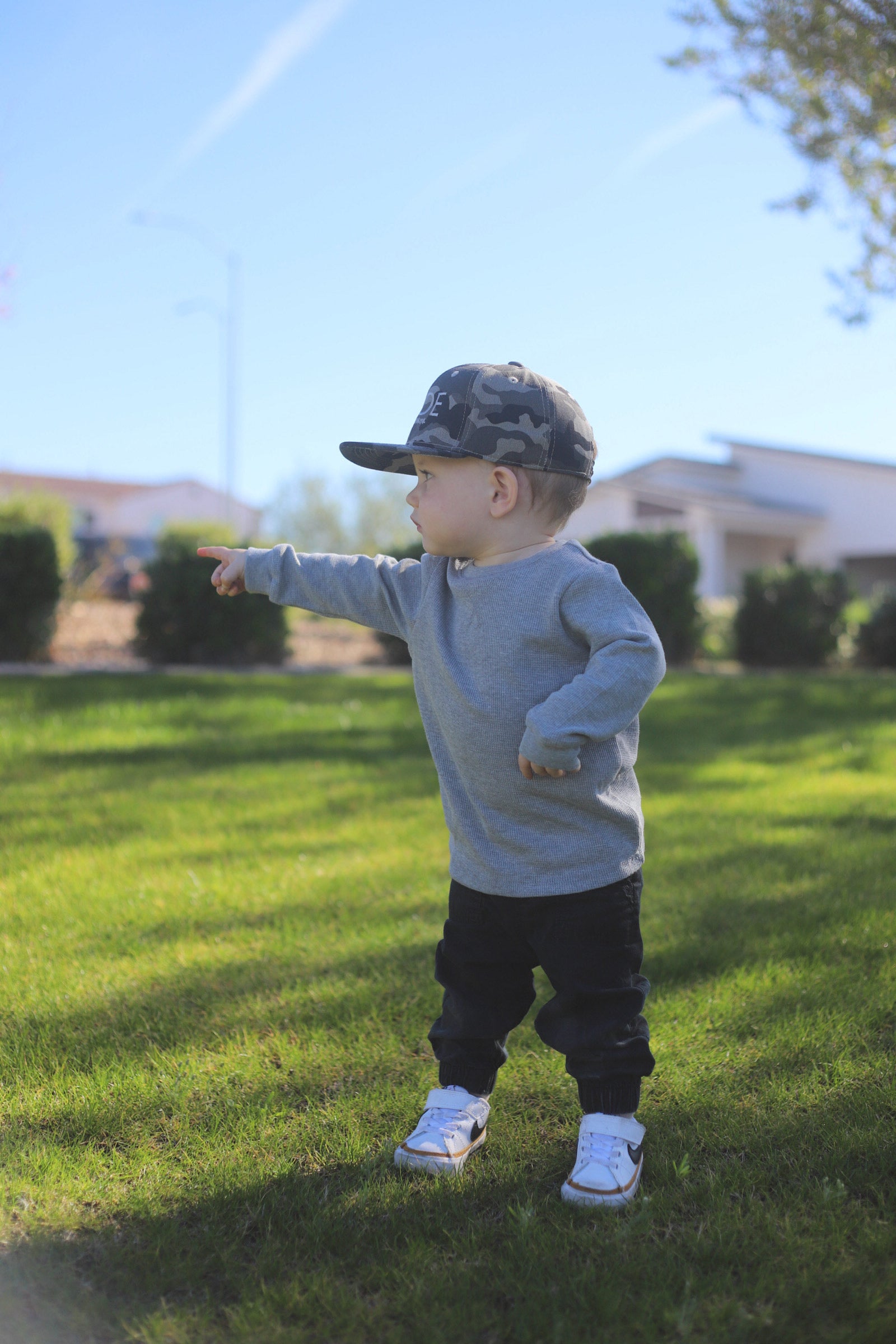 Toddler in camo flat bill boy hat and gray sweatshirt enjoying the outdoors.