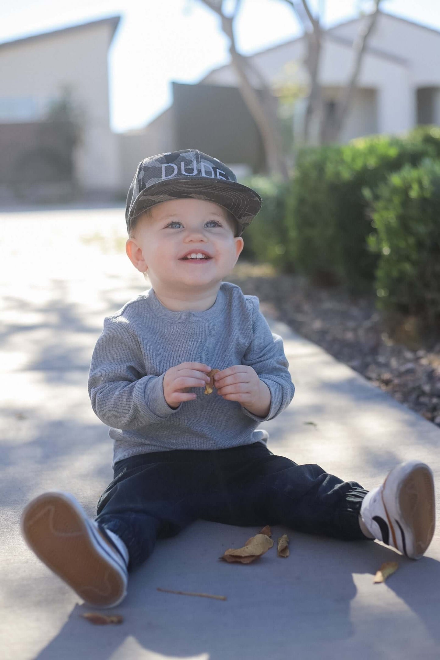 Baby boy smiling while sitting on the sidewalk wearing a boys toddler hat that says 'Dude'.