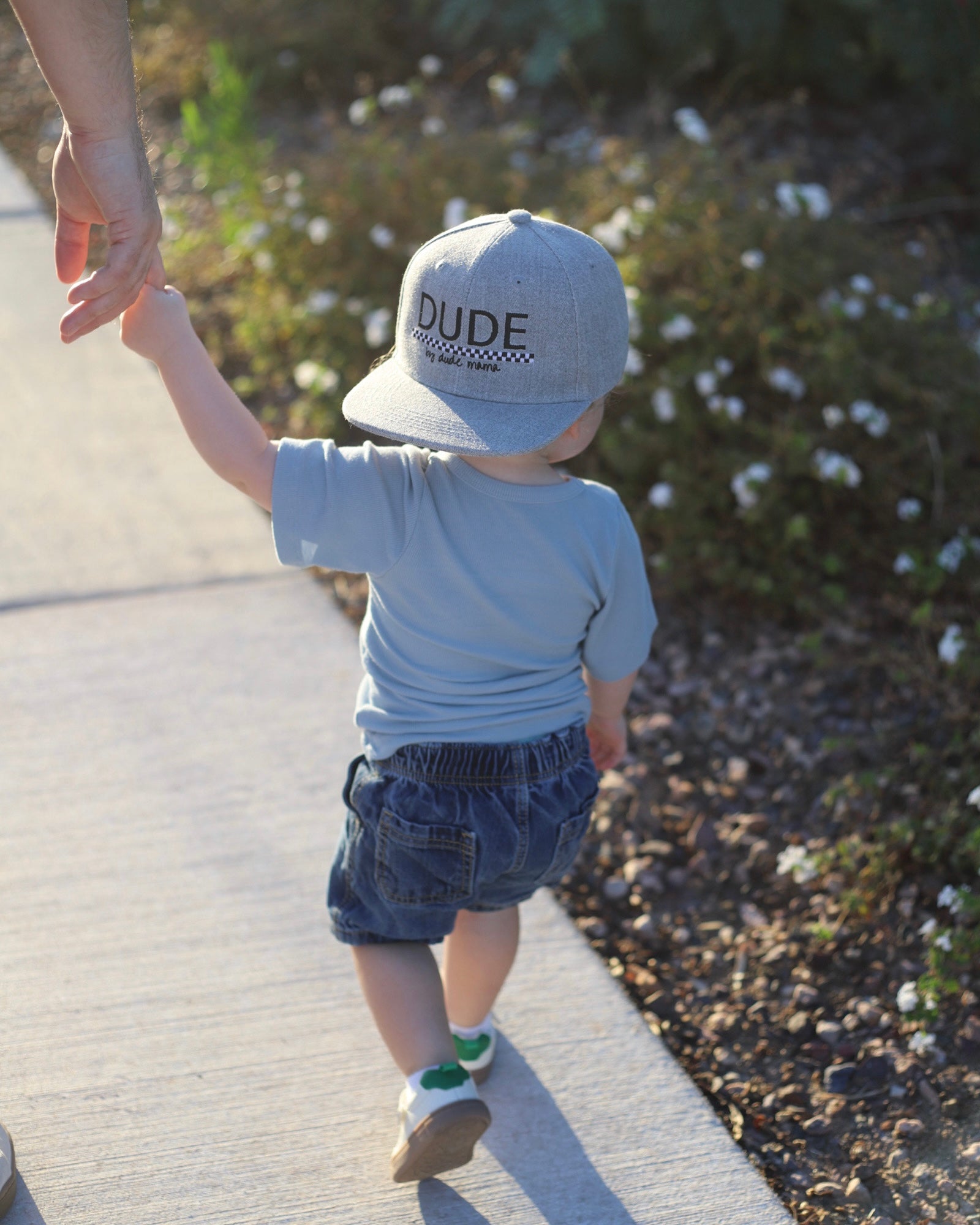 Young boy wearing a Light Gray Checkered Dude Hat while holding an adult's hand, enjoying a sunny day outdoors.