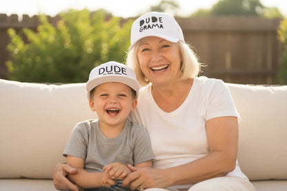 Grandma and grandson wearing matching white hats. Grandma's hat says 'Dude Grama' and her grandson's hat says 'Dude'.