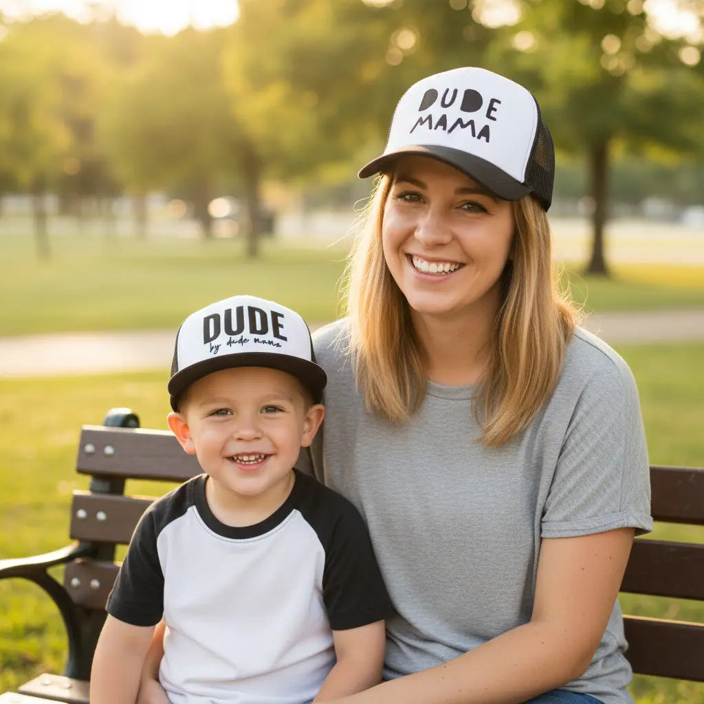 Mom and son sitting on a park bench wearing matching hats. Mom's hat says 'Dude Mama' and is a boy mom hat. Her son's hat is a kids hat and says 'Dude'. His hat is a flat brim and her hat is a curved brim. Makes a great mom and son matching outfit.