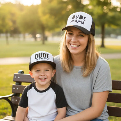Mom and son sitting on a park bench wearing matching hats. Mom's hat says 'Dude Mama' and is a boy mom hat. Her son's hat is a kids hat and says 'Dude'. His hat is a flat brim and her hat is a curved brim. Makes a great mom and son matching outfit.