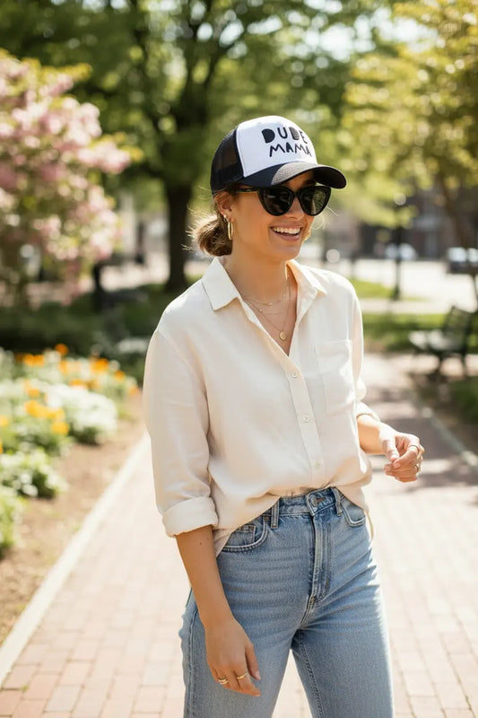 Mom smiling wearing a boy mom hat. It is black and white and says 'Dude Mama'. She is also wearing sunglasses and a light shirt in a park setting.